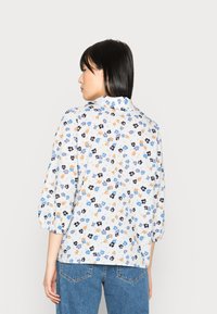 Blusa floral branca com padrões de flores azuis, laranja e pretas, mangas três quartos e um corte solto. Combinada com calças de cintura alta.