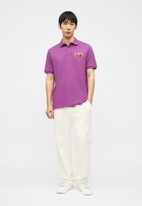 Άνδρας που φοράει μοβ πόλο με κεντήματα λουλουδιών, άσπρο παντελόνι και άσπρα αθλητικά παπούτσια, στέκεται με το ένα χέρι στην τσέπη.