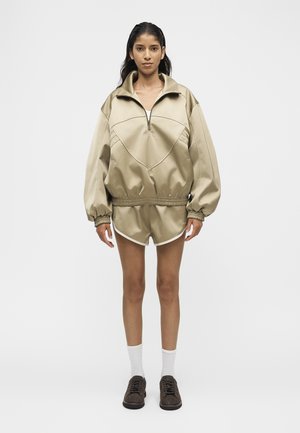 Žena s dlhými tmavými vlasmi má na sebe béžovú oversized bundu na zips a zodpovedajúce šortky s bielym lemom, biele ponožky a tmavé tenisky, stojí tvárou vpred.