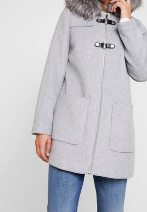 Kvinde iført en lysegrå uldfrakke med pelskrave, lynlåslukning, to sorte spænder og store forlommer over blå jeans.