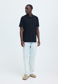 Polo noir à manches courtes avec col, associé à un jean bleu clair et des chaussures beige en slip-on. Design simple et décontracté.