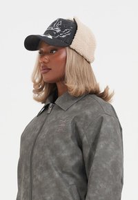 Black and beige cap with a textured wool lining and embroidered design. Worn with a gray faux leather zip jacket featuring a collar.