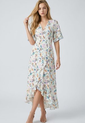 Mujer con vestido cruzado de flores con volante en el dobladillo, mangas cortas y tacones beige con tira en el tobillo, de pie frente a un fondo liso y claro.