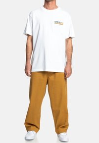 Bílé tričko s krátkým rukávem s malým zeleno-žlutým logem na hrudi, v kombinaci s volnými hořčicově žlutými kalhotami a bílými teniskami.