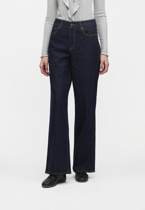 Femme portant un jean bleu foncé taille haute à jambes larges, un haut gris clair à manches longues et des chaussures noires, debout sur fond blanc.