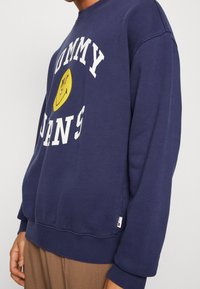 Tmavě modrá mikina vyrobená z bavlny, s velkým bílým potiskem "TOMMY JEANS" a žlutým logem usmívající se obličeje. Žebrované manžety a spodní lem.