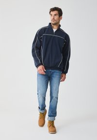 Sudadera de forro polar azul marino con ribete blanco, cuello con media cremallera, corte relajado; combinada con jeans azul claro y botas de cordones en color beige.