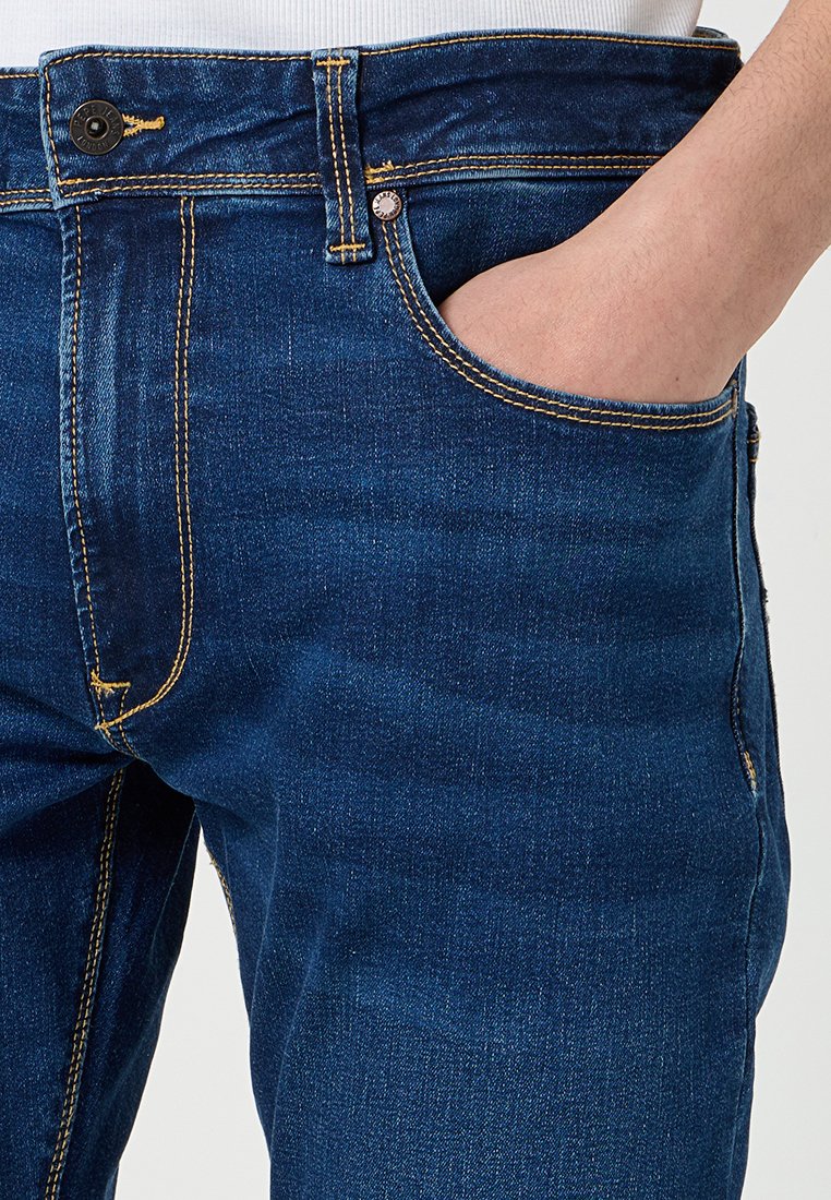 Hand in der Vordertasche von dunkelblauen Jeans aus Denim mit sichtbaren gelben Nähten und Metallschrauben sowie Nieten.