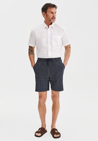 Biała koszula na krótki rękaw z guzikami i kieszonką, w połączeniu z ciemnoniebieskimi spodniami w paski. Lekki materiał; styl casualowy. Brązowe sandały.