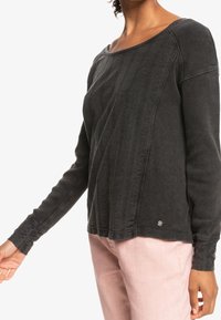 Vrouw in een donkergrijze textuur met lange mouwen en lichtroze broeken, getoond van de nek tot halverwege de bovenbenen, tegen een witte achtergrond.