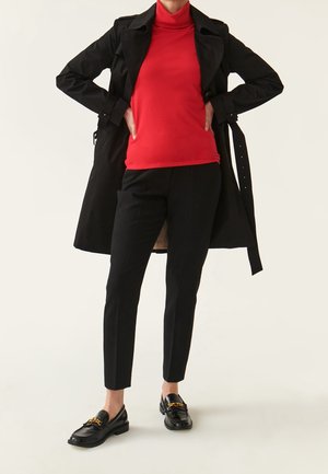 Manteau noir, pull rouge à col roulé, pantalons noirs ajustés et mocassins noirs avec un détail en chaîne dorée. Textures lisses, design épuré.