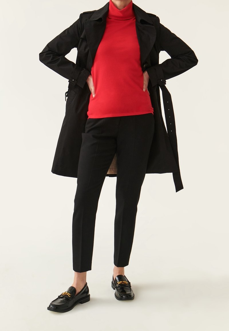 Czarny płaszcz trench, czerwony golf, dopasowane czarne spodnie i czarne loafersy z detalami w postaci złotego łańcuszka. Gładkie tekstury, minimalistyczny design.