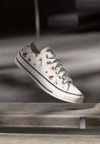White canvas sneakers with colorful floral embroidery, lace-up design, rubber toe cap, and black strip along the sole.