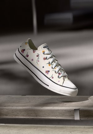Vita canvas sneakers med färgglad blommig broderi, snörningsdesign, gummitåskydd och svart rand längs sulan.