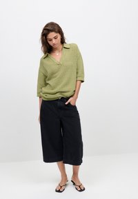 Maglione verde a maglia larga con scollo a V e maniche corte, abbinato a pantaloni neri a gamba larga e infradito nere. Design semplice e testurizzato.