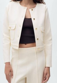 Elfenbeinfarbene kurze Jacke mit goldenen Knöpfen, zwei Brusttaschen und rundem Ausschnitt, kombiniert mit einem braunen Rippentop und passenden Hosen.