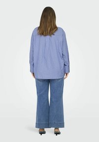 Lichtblauw en wit gestreepte oversized blouse met lange mouwen, gecombineerd met brede blauwe denim broek en zwarte hakken.