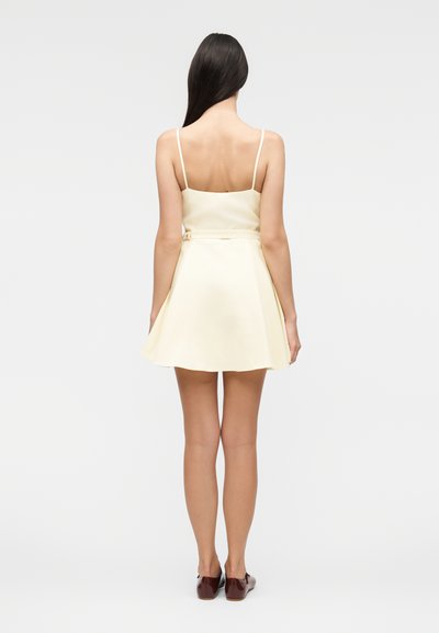 Γυναίκα με μακριά σκούρα μαλλιά που φοράει ένα αμάνικο, ανοιχτό κίτρινο φόρεμα και καφέ ίσια παπούτσια, στέκεται με την πλάτη προς τον θεατή σε μονόχρωμο φόντο.