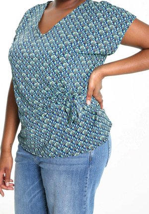 Femme portant un chemisier bleu à manches courtes à motifs avec un nœud sur le côté et un jean bleu clair, la main posée sur la hanche, sur fond blanc.