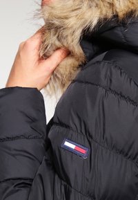 Czarna kurtka puchowa z kapturem wykończonym futrem. Posiada pikowane sekcje, naszywkę z logo w kolorach czerwonym, białym i niebieskim oraz gładką fakturę.