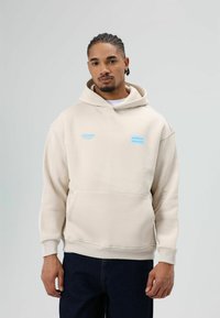 Beige hoodie met een voorzak, voorzien van blauwe opdruk met de tekst "STRONGER" en "chance." De stof lijkt zacht met geribbelde manchetten en zoom.