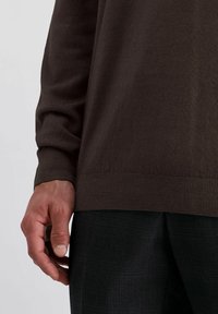 Nahaufnahme der linken Hand einer Person, die an ihrer Seite ruht, und die einen dunkelbraunen Pullover und schwarze strukturierte Hosen trägt.