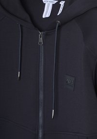 Mornarica zip-up dukserica s glatkom teksturom, bočnim džepovima i malim gumenim logom. Ima metalni zatvarač i akcentne vezice.