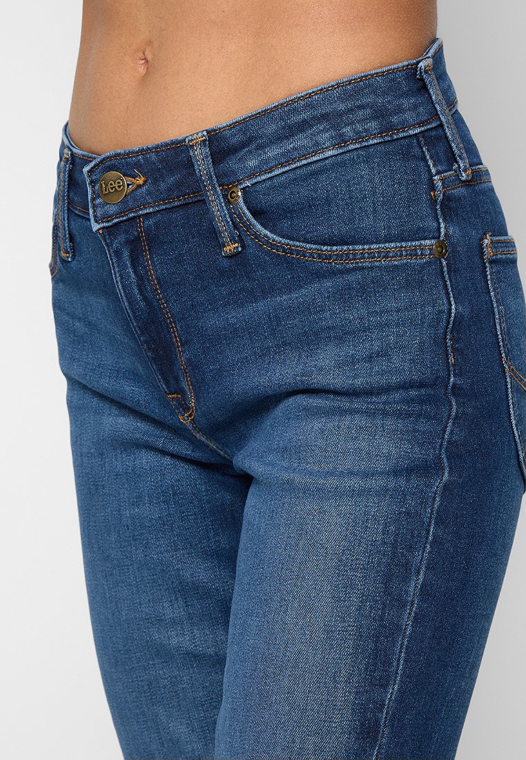 Gros plan sur le bas-ventre portant un jean bleu de la marque Lee avec des coutures oranges et des poches avant, montrant une partie de peau nue au-dessus de la taille.