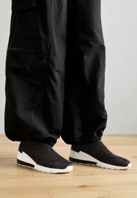 Svarta slip-on sneakers med en texturerad ovandel, vit sula och svarta accenter. Bärs med löst sittande svarta byxor på ett trägolv.