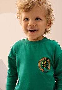 Felpa verde in cotone con un logo rotondo che raffigura alberi e la scritta "Adventure Awaits" in arancione, su un tessuto strutturato.