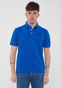 Polo blu in cotone, con colletto, tre bottoni, maniche corte e un piccolo patch logo nero sul petto. Indossato con jeans.