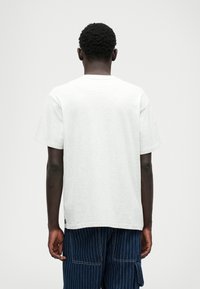 Svijetlosiva pamučna majica s opuštenim krojem, okruglim izrezom, kratkim rukavima i bez vidljivih uzoraka ili grafike, u kombinaciji s tamnim prugastim hlačama.