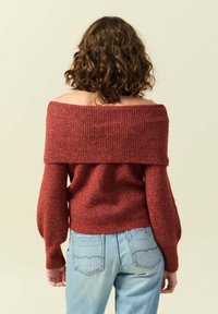 Maglione a spalle scoperte di colore bordeaux in maglia, con coste testurizzate, maniche voluminose e un corpo aderente, abbinato a jeans azzurri chiari.