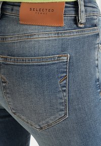 Ljusblå denimjeans med en närbild bakifrån, med en läderlogotyp dekaler och en enda bakficka med sömnadsdetaljer.