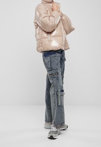 Kurtka puchowa w błyszczącej, jasnej beżu, z wysokim kołnierzem i pikowanym wzorem. W połączeniu z niebieskimi dżinsami cargo w stylu patchwork i szarymi trampkami.