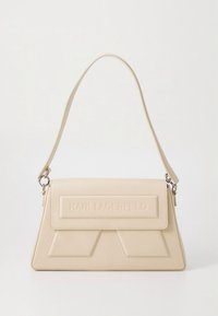 Bēša ādas rokas soma ar strukturētu dizainu, reljefa logo atlokā un vienu plecu siksnu. Gluda tekstūra, asas līnijas.