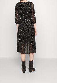 Robe noire avec un motif tacheté doré, manches longues semi-transparentes, taille marquée et ourlet asymétrique. Associée à des bottines noires.