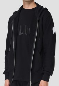 Felpa nera con zip, dal tessuto liscio, caratterizzata da una vestibilità comoda, tasche a marsupio e dettagli in bianco sulle maniche.