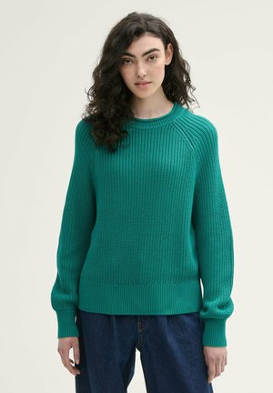 Türkiser Strickpullover mit gerippter Textur, Rundhalsausschnitt und leicht gepufften langen Ärmeln. Kombiniert mit dunkelblauen Jeans.