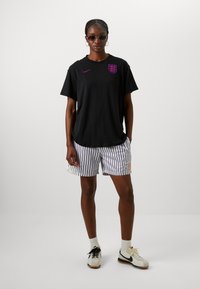Czarny, oversize'owy t-shirt z fioletowym logo Nike i godłem Anglii, dobrany do białych i czarnych pasiastych szortów z pomarańczowymi detalami.
