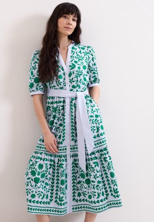 Frau mit langen dunklen Haaren, die ein weißes Midikleid mit grünen Blumenmustern und einem gebundenen weißen Gürtel trägt, steht vor einer schlichten weißen Wand.