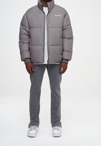 Doudoune grise avec un col montant, fermeture éclair à l'avant et détail de logo. Portée sur un T-shirt blanc, accompagnée de pantalons gris et de baskets blanches.