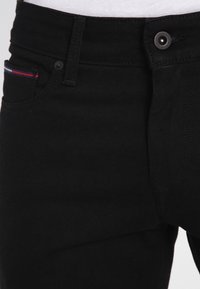 Black denim jeans featuring a brass button, subtle stitching, and a small multi-colored accent on the front pocket.
