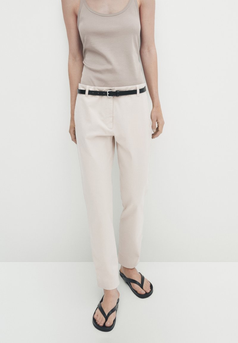 Beige tanktop, lichtgekleurde broek met een zwarte riem en zwarte teenslippers. Glad stof, rechte snit en minimale details.