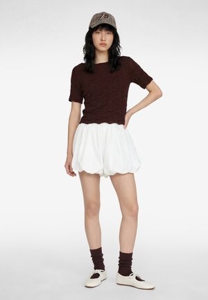 Junge Frau steht mit einer Hand in der Hüfte, trägt ein braunes, strukturiertes Kurzarmoberteil, weiße, gepuffte Shorts, braune Socken, weiße Schuhe und eine graue Mütze.