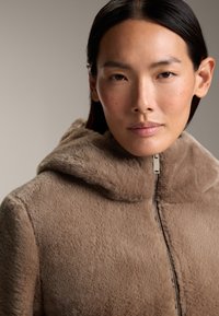 Brun fuskpälsjacka med huva, med dragkedjestängning och mjuk textur. Designen är slät utan synliga mönster eller detaljer.