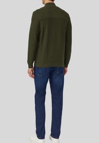 Olivengroen gebreid vest met een geribbelde kraag en bovenrug, met een gestructureerd ontwerp, gecombineerd met blauwe rechte jeans en witte sneakers.