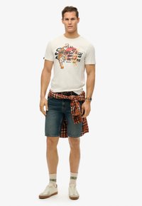 White graphic t-shirt featuring a tiger design, paired with blue denim shorts. A red and black checkered shirt is tied at the waist. White sneakers complete the look.