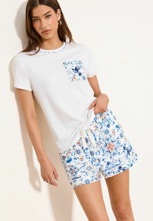 Naine, kes kannab valget T-särki sinise linnu taskuga ja sobiva lillmustrilise sinise ja oranži lühikeste pükstega, seisab valge seina taustal.