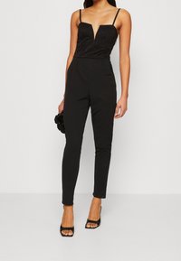 Mono negro con un escote de encaje profundo, tirantes finos, silueta ajustada y piernas acampanadas. Combinado con tacones negros con tiras y un bolso de mano.